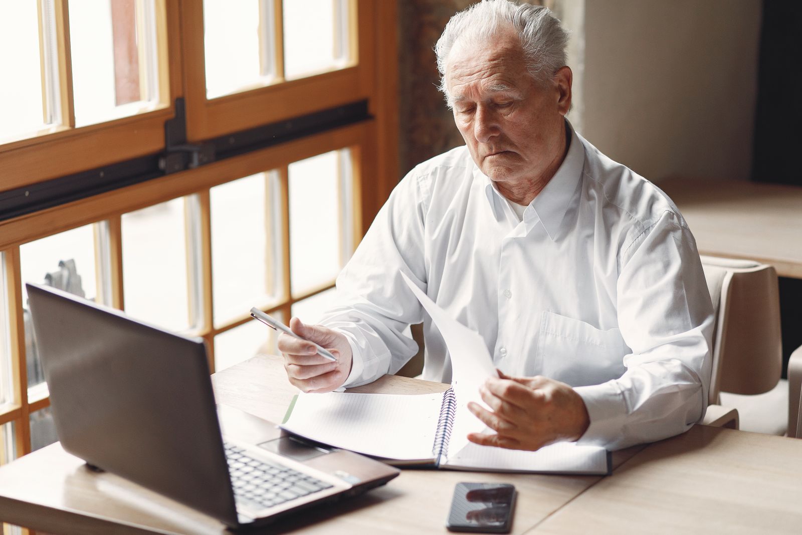 An older man reviewing notes beside a laptop at his desk