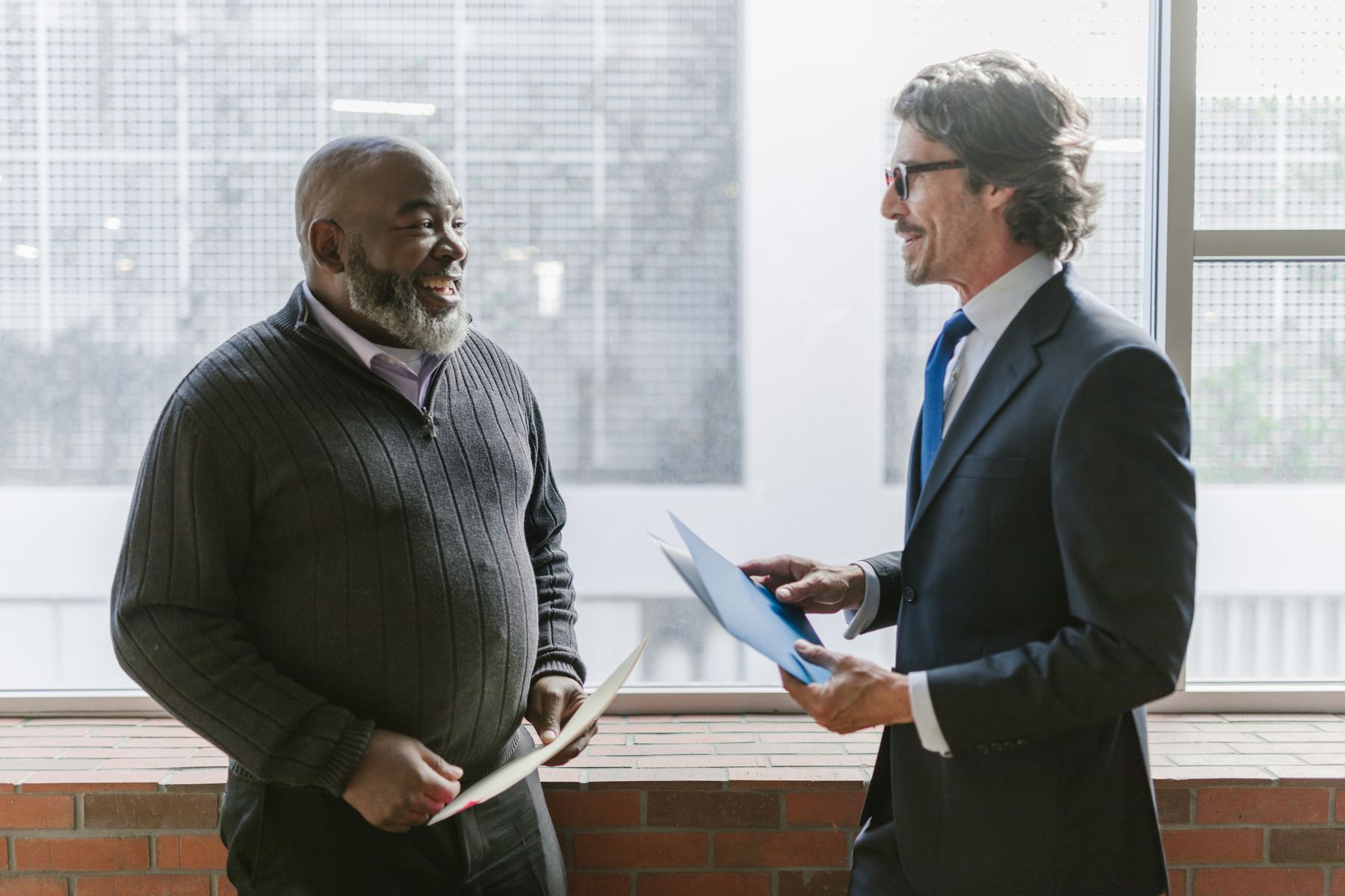 Two men discussing documents beside an office window